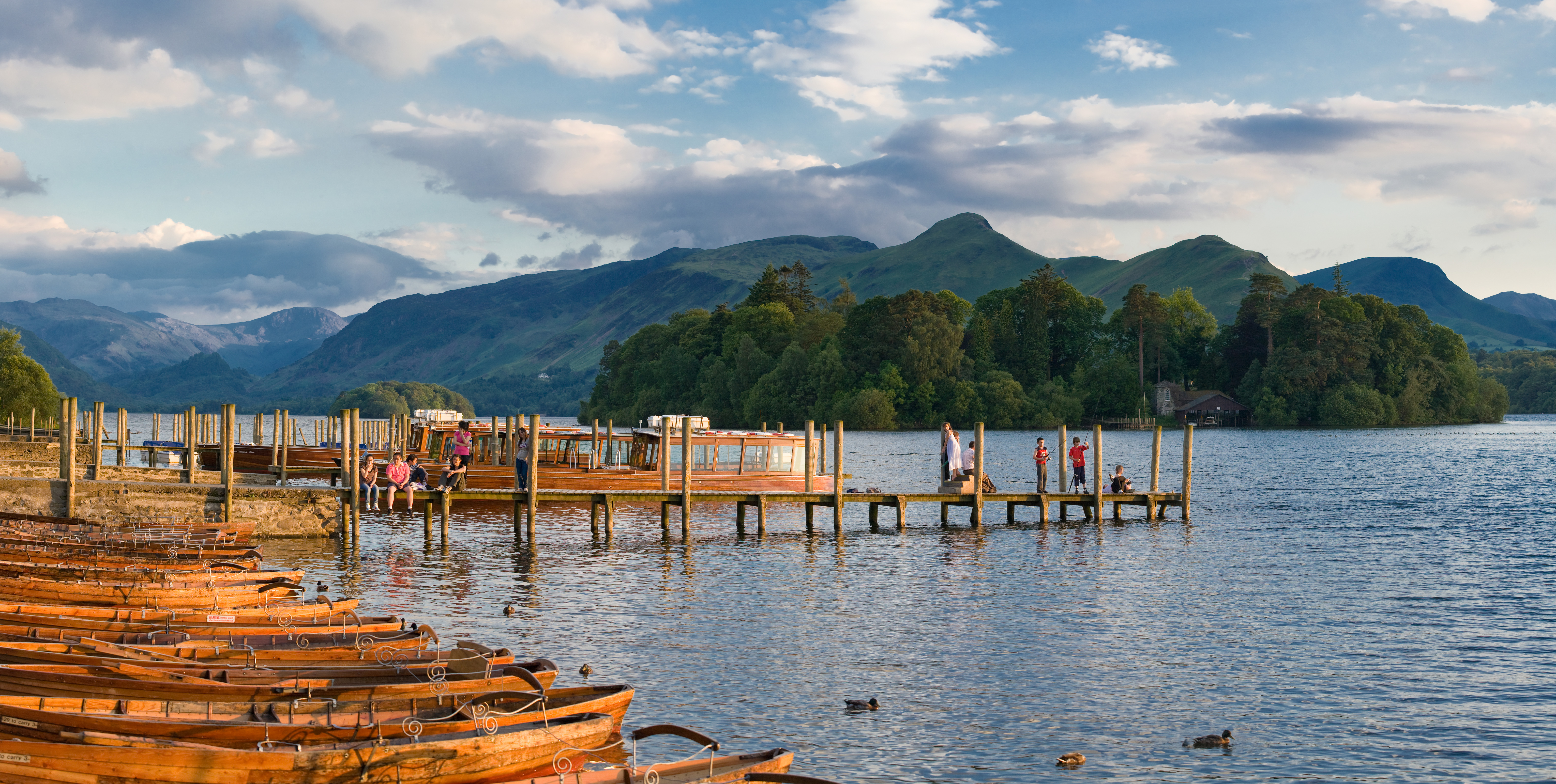 Calm lake scene with wooden boats docked near a pier. People stand and sit on the pier, with forested hills and mountains in the background under a blue sky