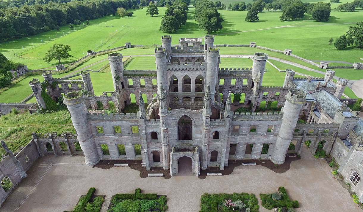 Aerial view of a partial medieval castle ruin with tall, cylindrical towers and arched windows, surrounded by lush green lawns and trees.