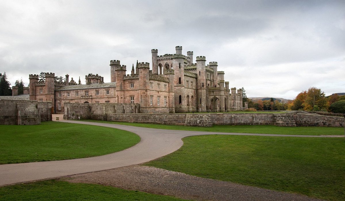 A grand stone castle with towers and battlements under a cloudy sky, surrounded by lush green lawns and trees with autumn foliage in the distance
