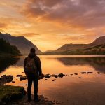 Person with backpack admires scenic lakeside sunset in mountainous landscape